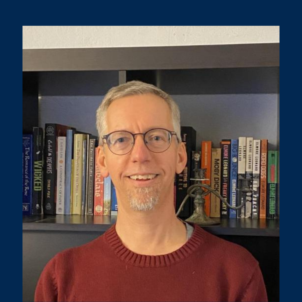 Image of Dr. Carl Whithaus smiling in front of a bookshelf against a dark blue background.